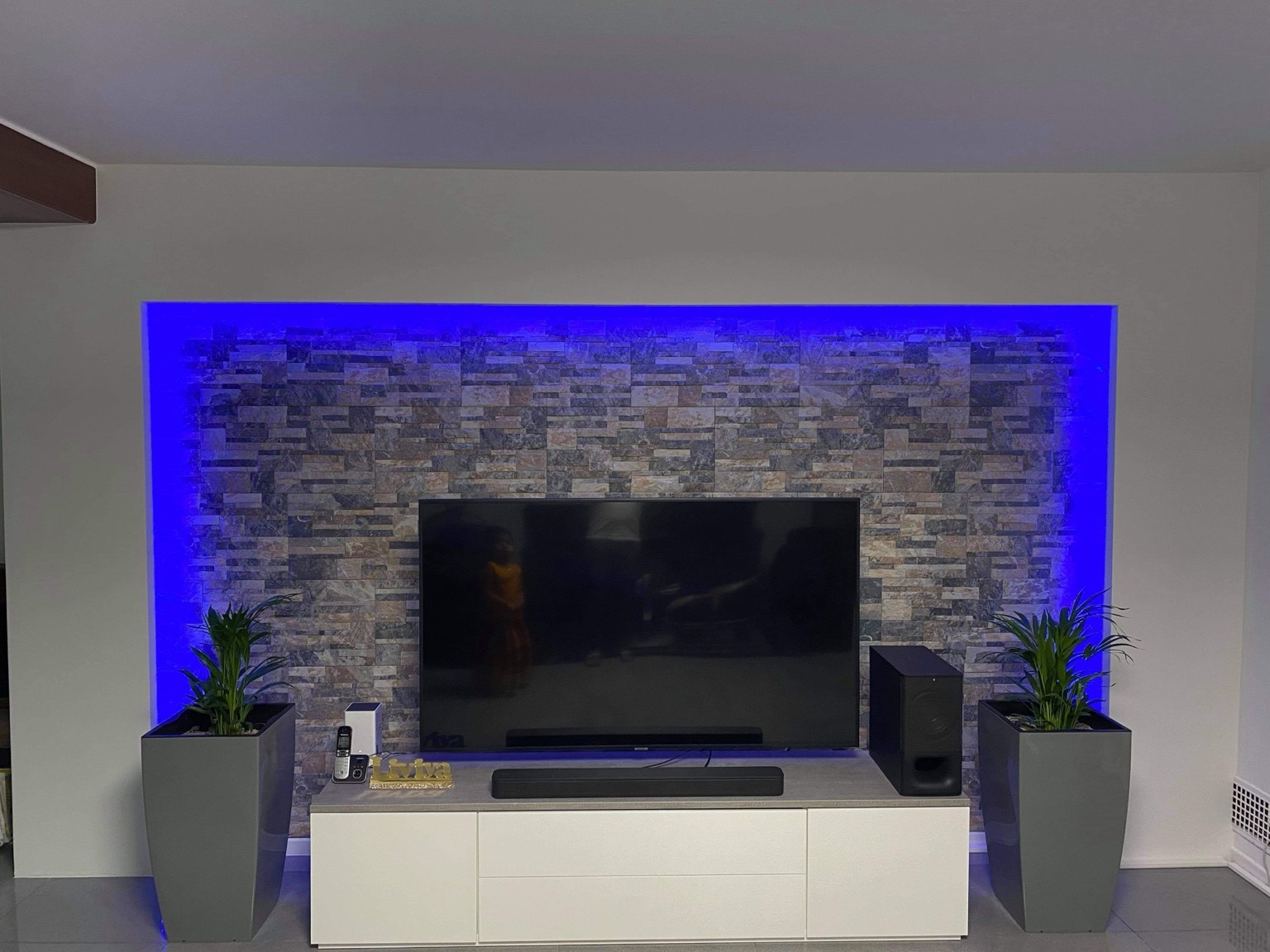 Weisse Einfassung um eine TV-Wand in Steinoptik mit umlaufender blauer Beleuchtung.